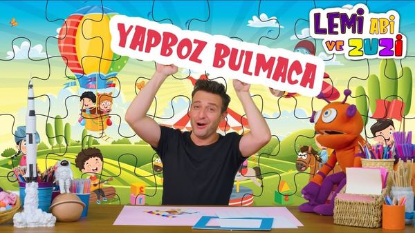 Kaydırmalı Yap-Boz video kapağı: sunucu Yapboz Bulmaca yazılı pankart tutuyor, arkada renkli yapboz sahnesi